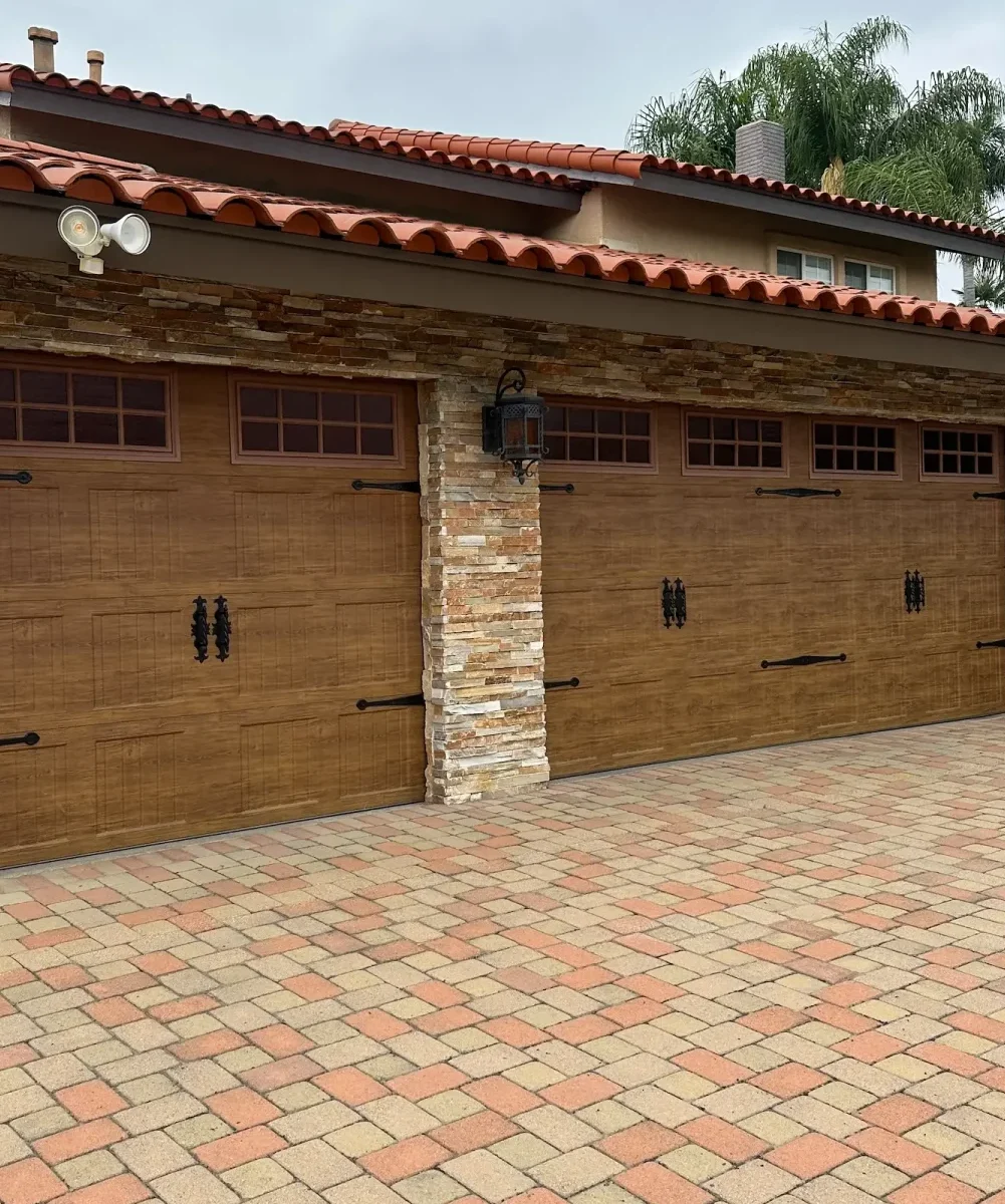 A house with two wooden garage doors, a stone pillar between them, a lantern-style light, red-tiled roof, and a paved driveway with tan, red, and brown bricks. Carlos Garage Door Repair provides expert garage door service for this stylish home.