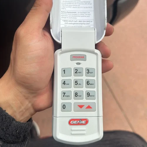 A hand holds a Genie garage door keypad with the cover flipped open, revealing a numeric keypad and “PROGRAM” button at the top. The Genie logo is visible near the bottom—perfect for those seeking reliable garage door repair Orange County services.