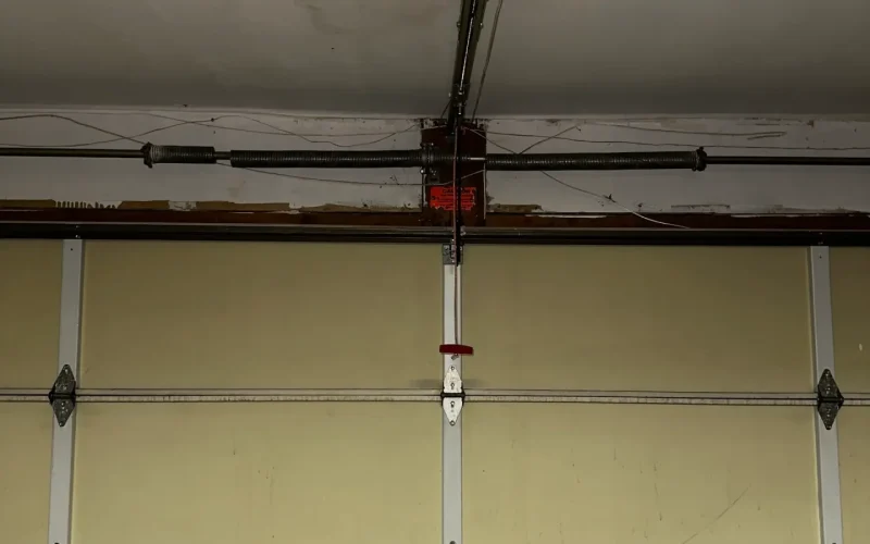 A beige garage door with metal hinges is shown closed. Above it, a garage door opener mechanism with a red emergency release handle hangs from the rail near the ceiling, which has visible water stains—ideal for a garage door repair service.