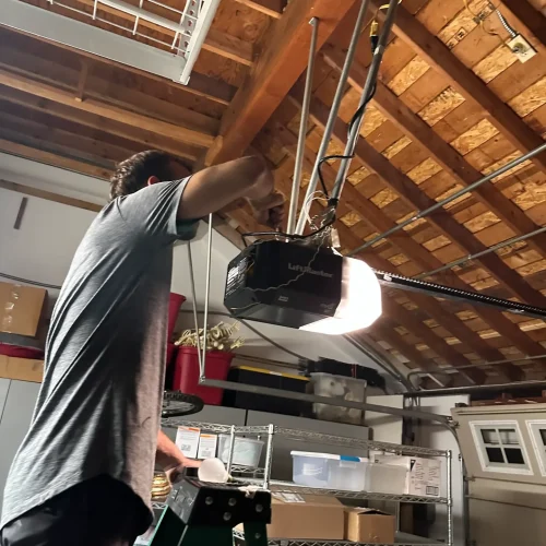 A person standing on a ladder inspects a garage door opener attached to the ceiling in a garage with exposed wooden beams and storage shelves, providing expert garage door repair.