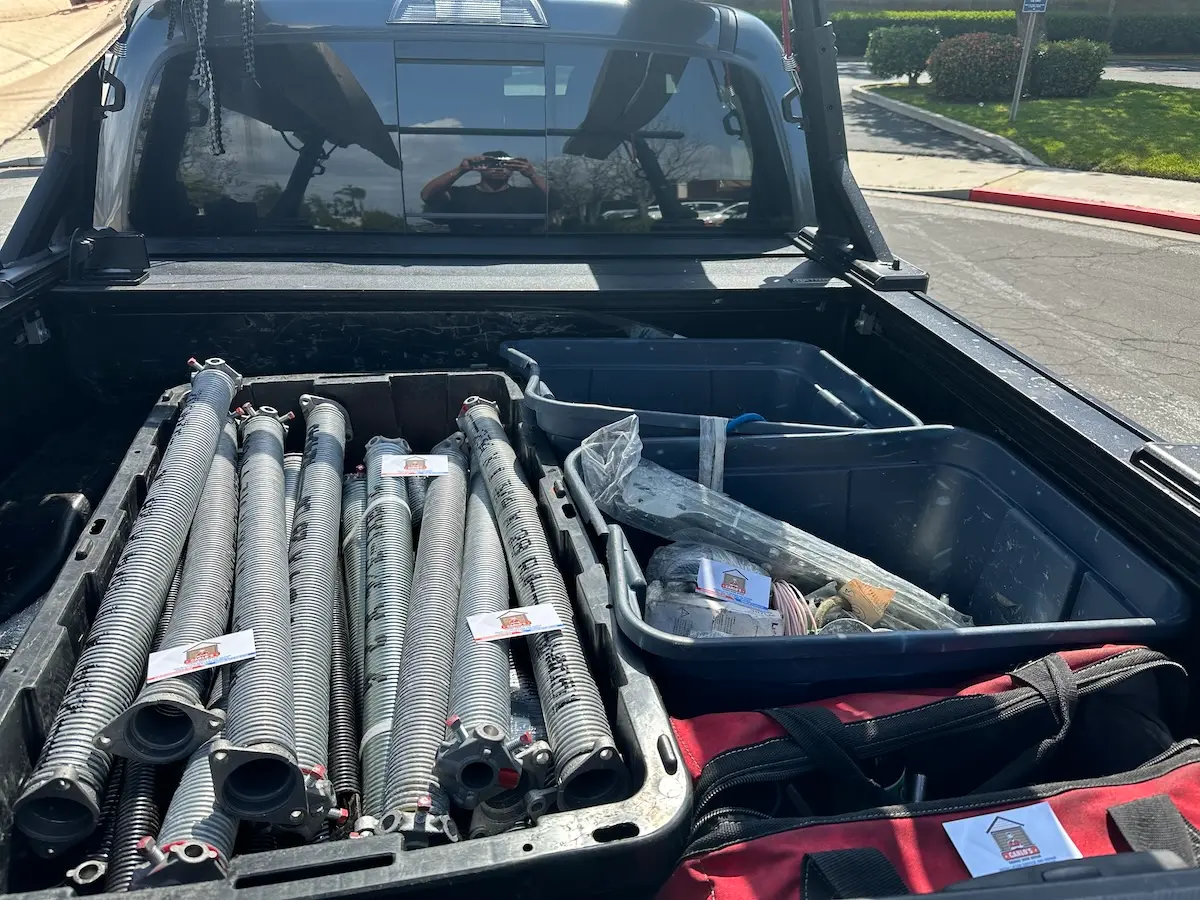 The bed of a pickup truck from Carlos Garage Door Repair holds several long metal garage door springs, a toolbox, and a red-and-black tool bag. The truck is parked on a sunny residential street in Orange County.